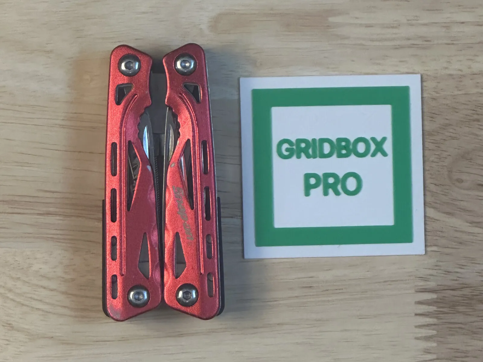 A red multitool lying flat next to the printed Gridbox Pro scaler tile, shot from straight above